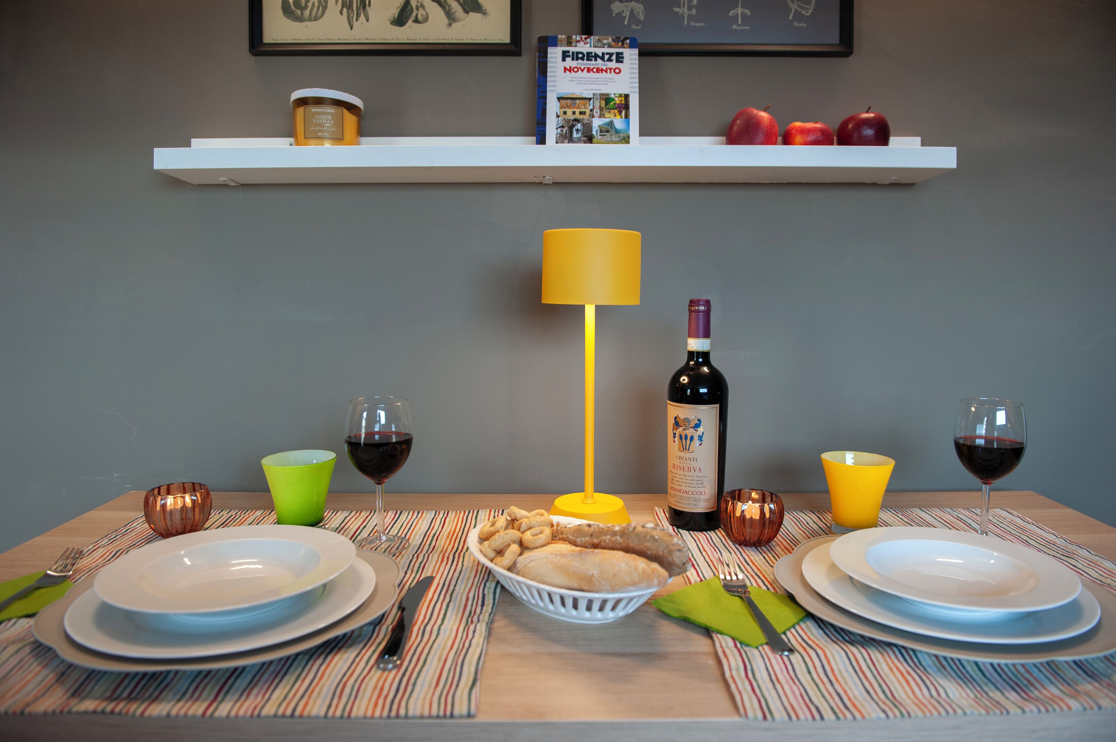 Una mesa de comedor dispuesta con platos, cubiertos, una botella de vino, copas y una cesta de aperitivos.