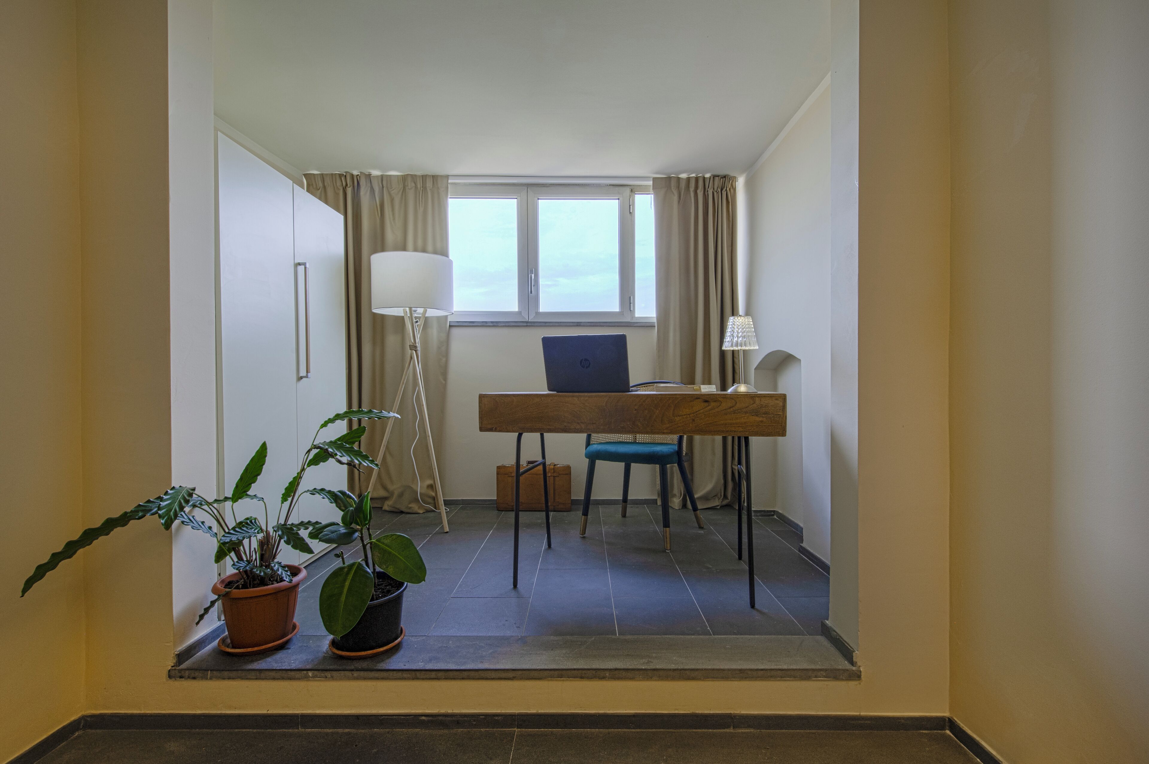 Ein kleines Zimmer mit Schreibtisch, Stuhl, zwei Topfpflanzen, einer Lampe und einem Koffer.