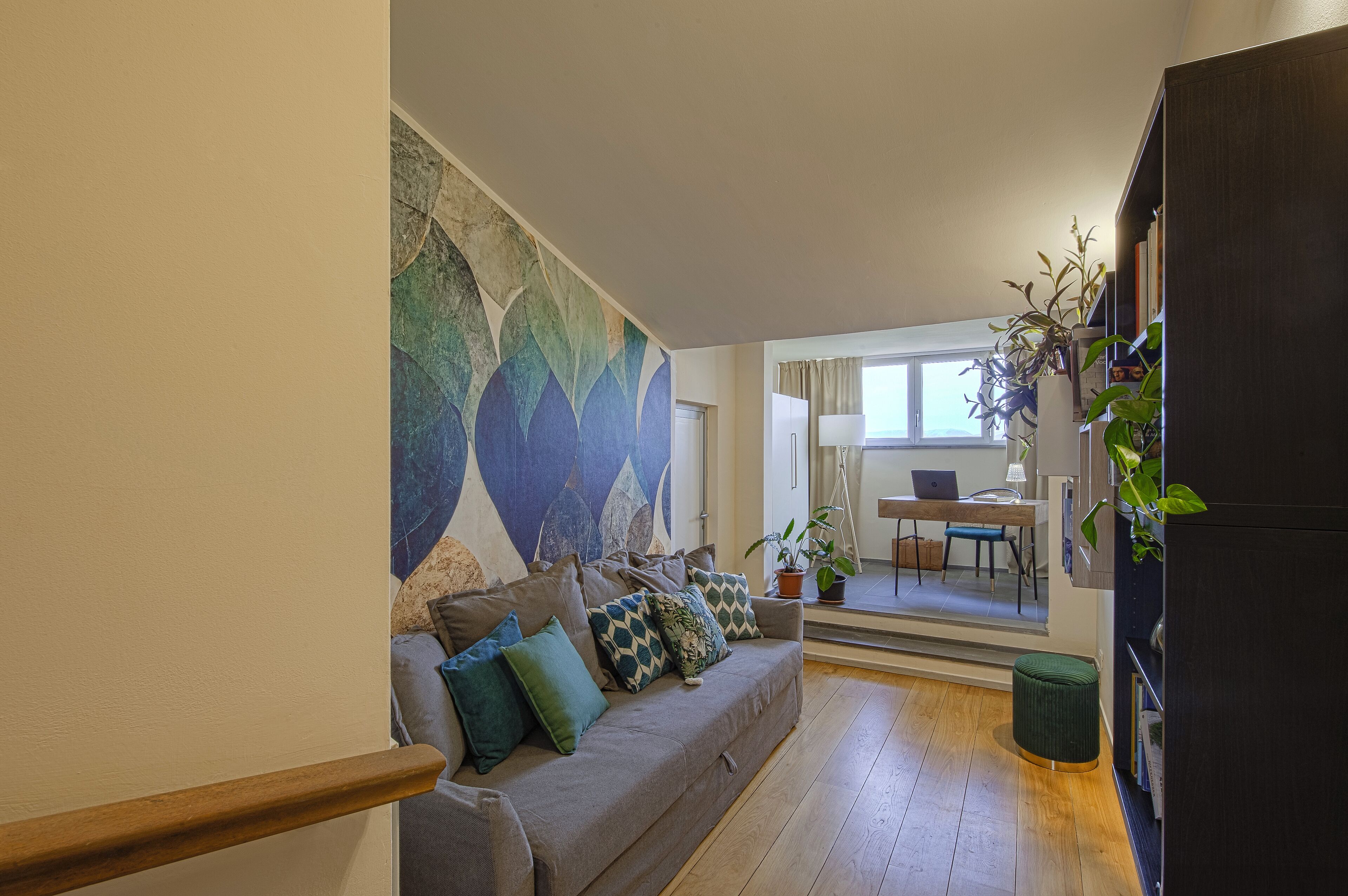 A modern living room with a grey sofa, a colorful abstract wall mural, wooden flooring, and a staircase.