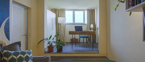 A room with a wooden floor, a sofa with patterned cushions, a desk with a laptop, a standing lamp, and a potted plant.