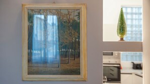 Ein gerahmtes Gemälde einer Waldszene an einer Wand, ein Küchenbereich mit Geräten und ein Fenster mit einem grünen Gegenstand im Fensterbrett.