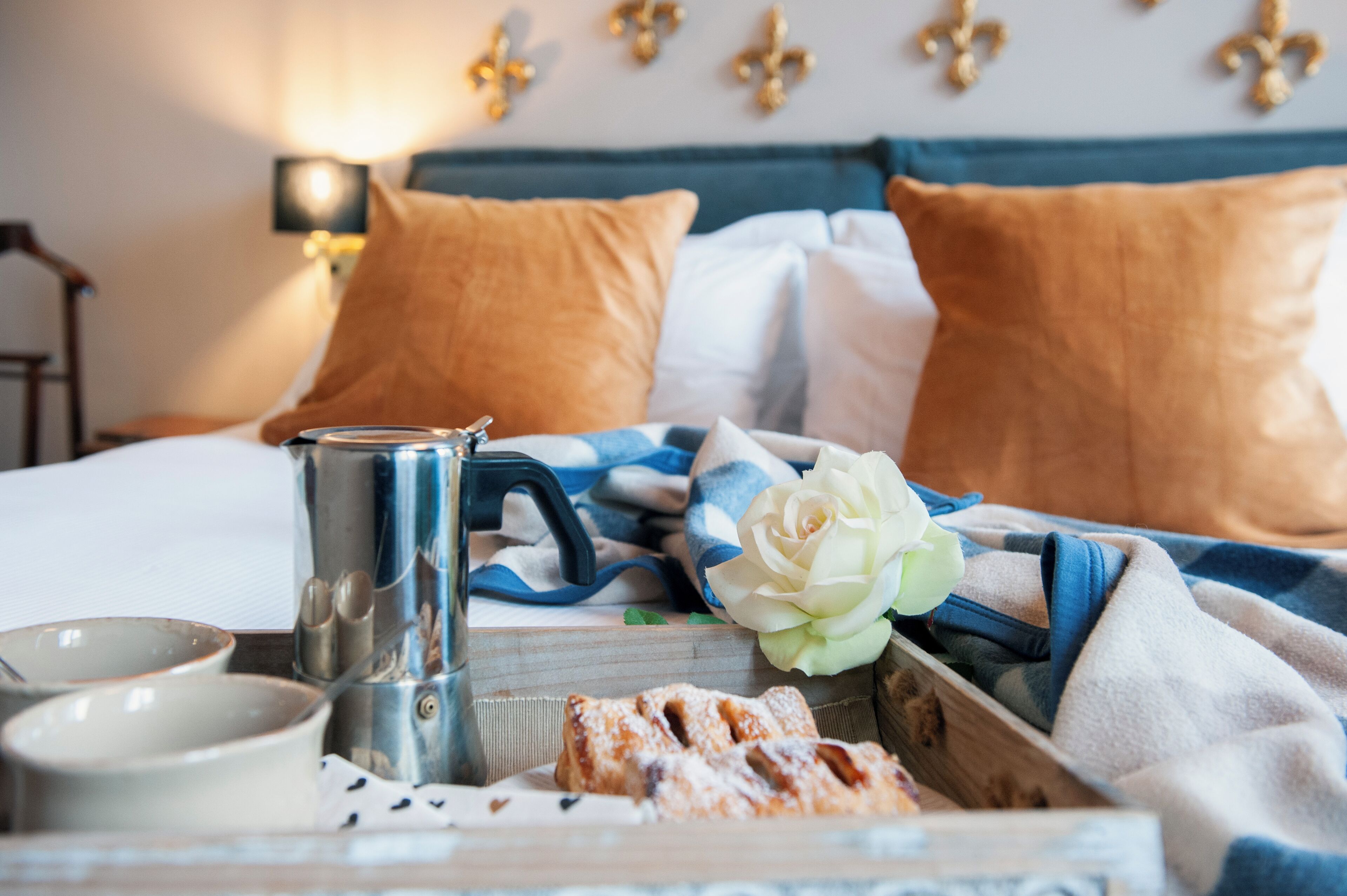 A neatly arranged breakfast tray with a coffee pot, a bowl, and pastries on a bed with a blue headboard and decorative pillows.