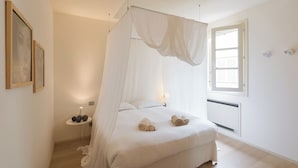 A bedroom with a canopy bed, bedside table with decorative items, and a window with blinds. - Boboli - Flo Apartments (Florence)