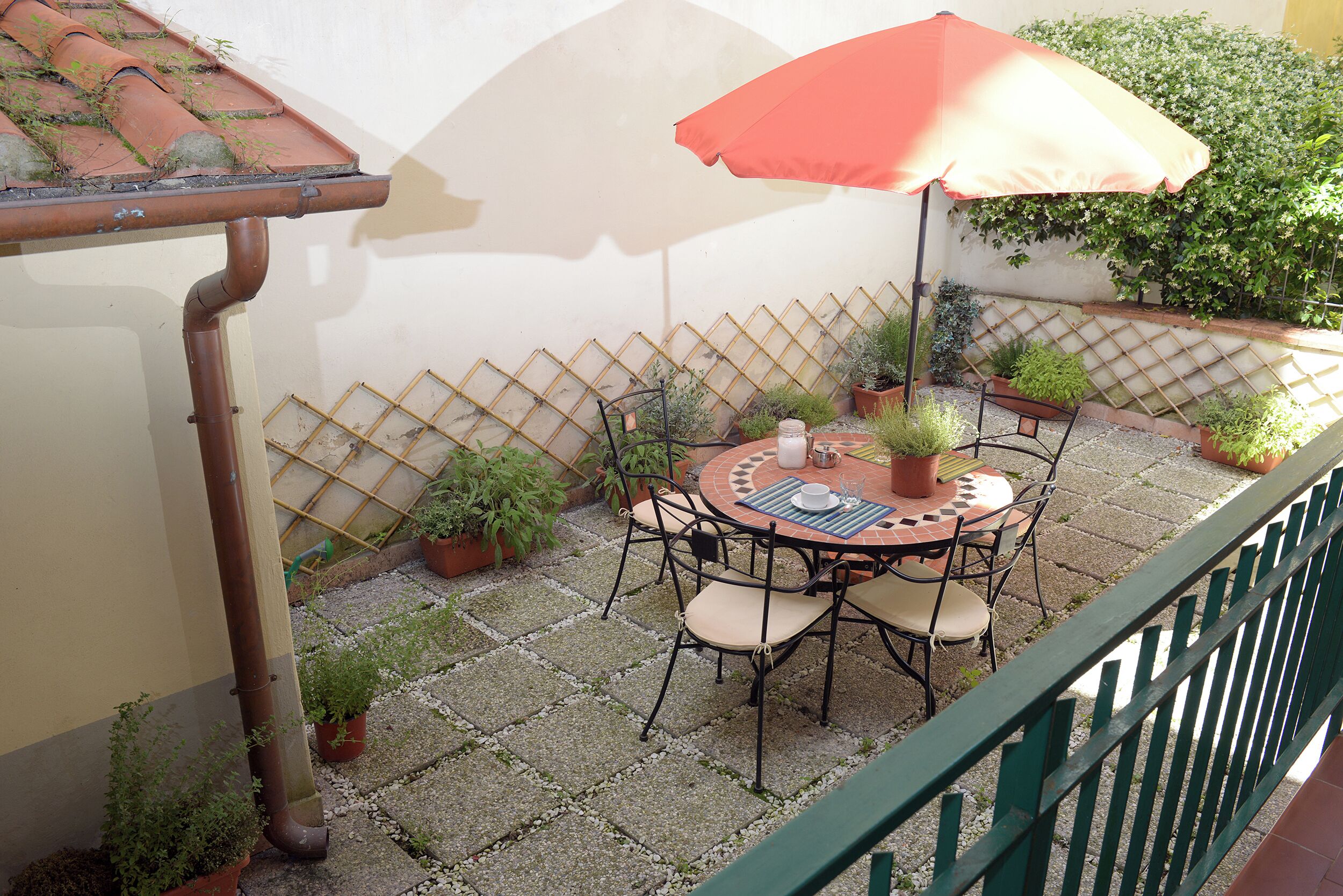 A patio with a round table and chairs, surrounded by potted plants and a red umbrella.