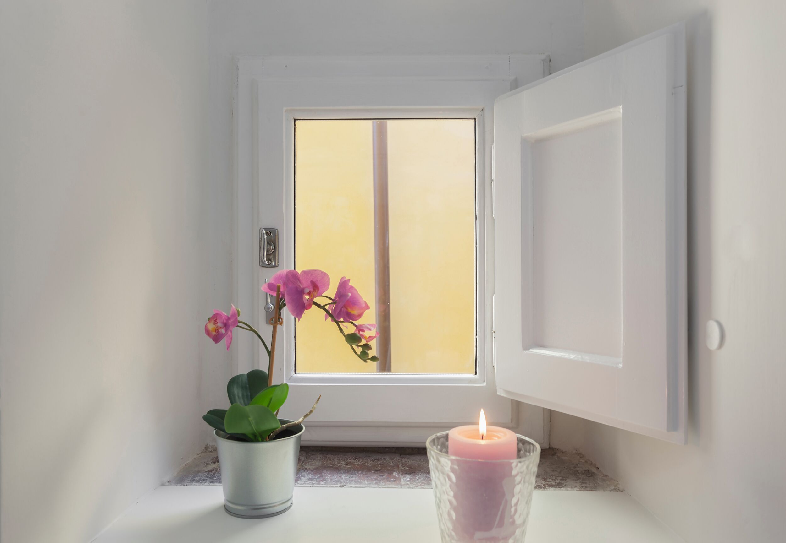 Ein kleines Fenster mit einer weißen Tür, einem Blumentopf und einer brennenden Kerze auf einem Fensterbrett.