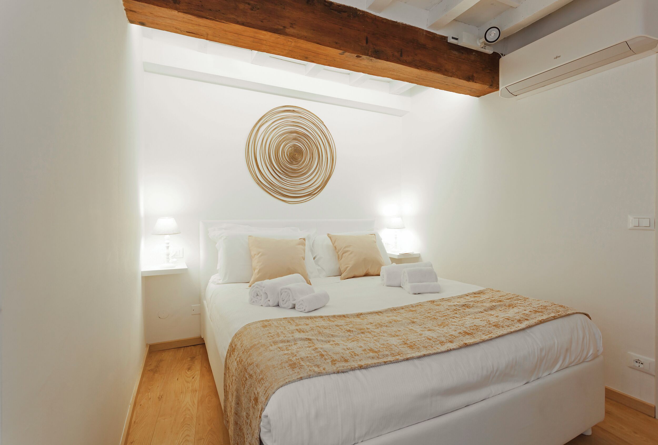 A bedroom with a bed, bedside lamps, a circular wall decoration, and a wooden ceiling beam.