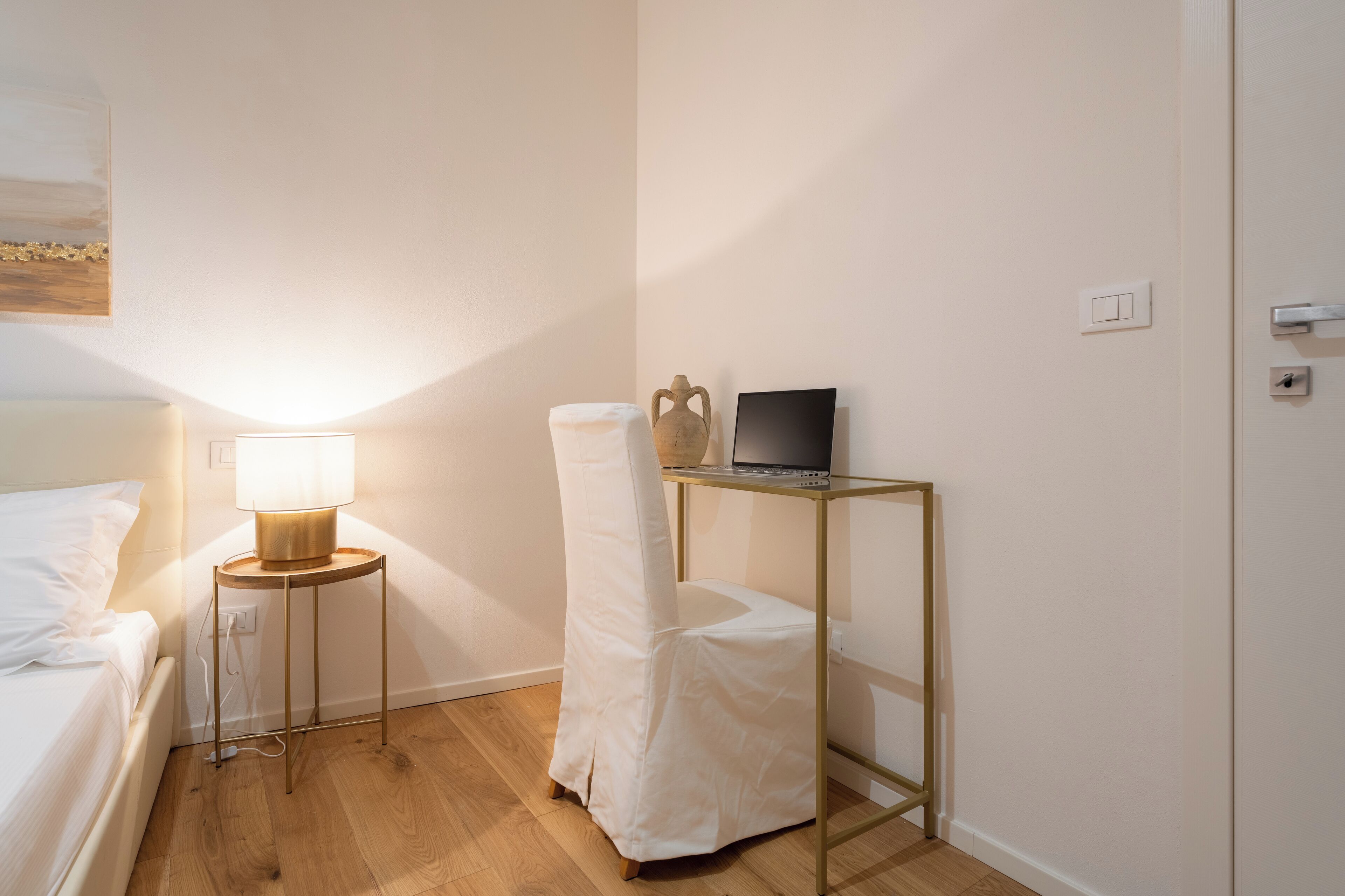 Ein ordentlich eingerichtetes Schlafzimmer mit einem Bett, einer Nachttischlampe, einem Laptop auf einem kleinen Tisch und einem Sessel mit weißem Bezug.