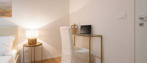 Ein ordentlich eingerichtetes Schlafzimmer mit einem Bett, einer Nachttischlampe, einem Laptop auf einem kleinen Tisch und einem Sessel mit weißem Bezug.