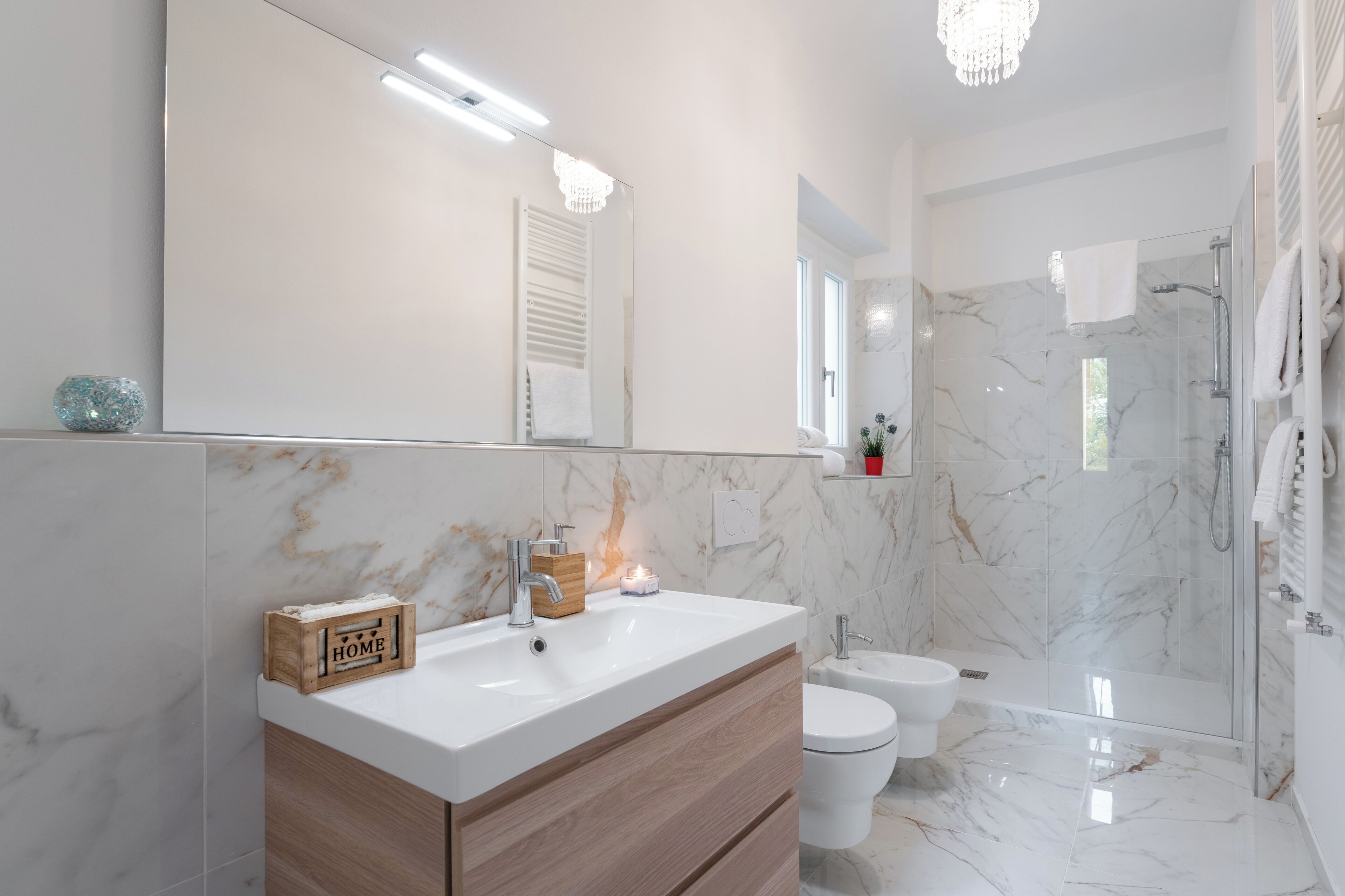 Une salle de bain moderne avec un grand miroir, des carreaux de marbre blanc, un meuble-lavabo en bois, des toilettes et une douche séparée par des parois en verre.