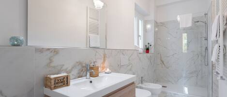 Une salle de bain moderne avec un grand miroir, des carreaux de marbre blanc, un meuble-lavabo en bois, des toilettes et une douche séparée par des parois en verre.