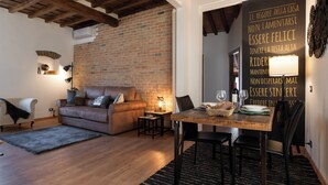 A cozy living room with a brick wall, a wooden ceiling, a leather sofa, a small table with a lamp, and a dining area with a wooden table and chairs. - Santa Trinita - Flo Apartments (Florence)