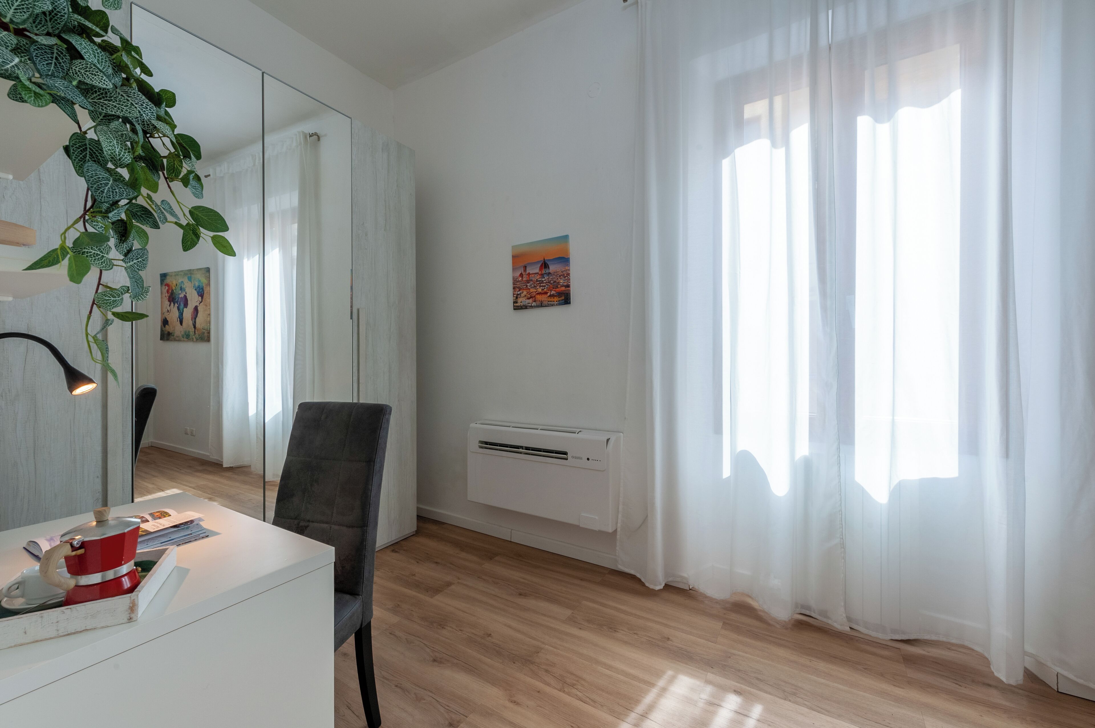 A modern room with a wooden floor, a white desk with a red kettle, a chair, a large mirror, and a radiator.