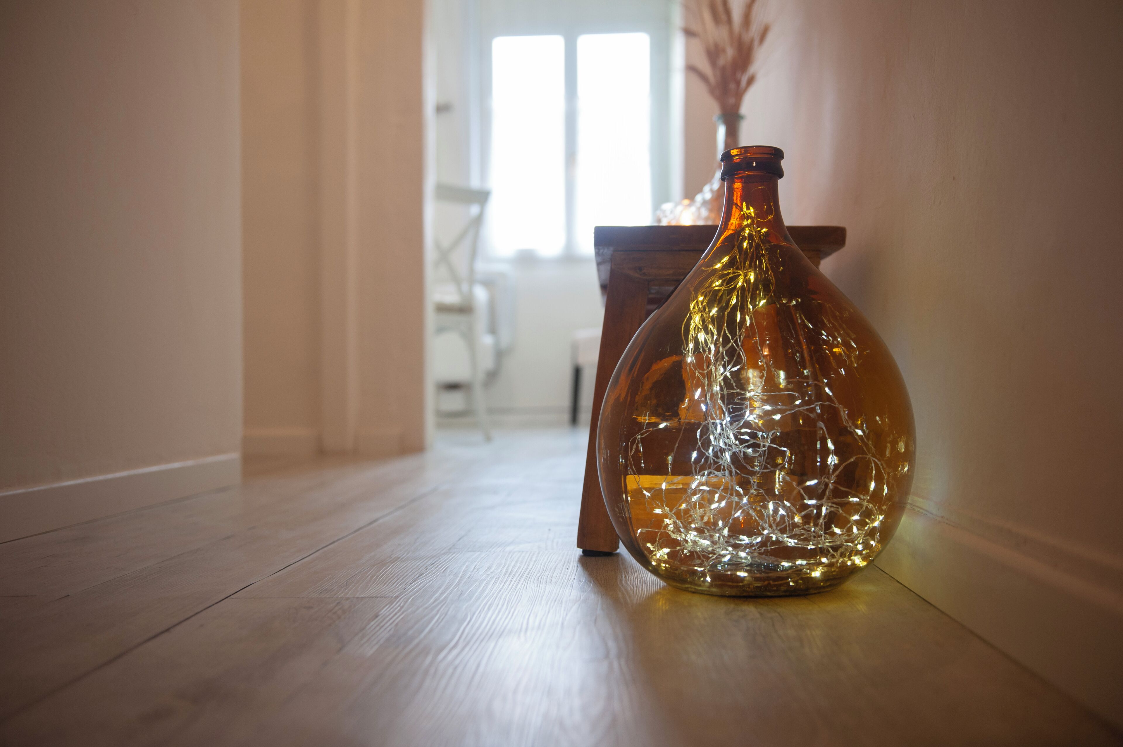 Une bouteille en verre transparent, dotée d’une surface texturée, posée sur un plancher en bois. On aperçoit une fenêtre et une chaise à l’arrière-plan.