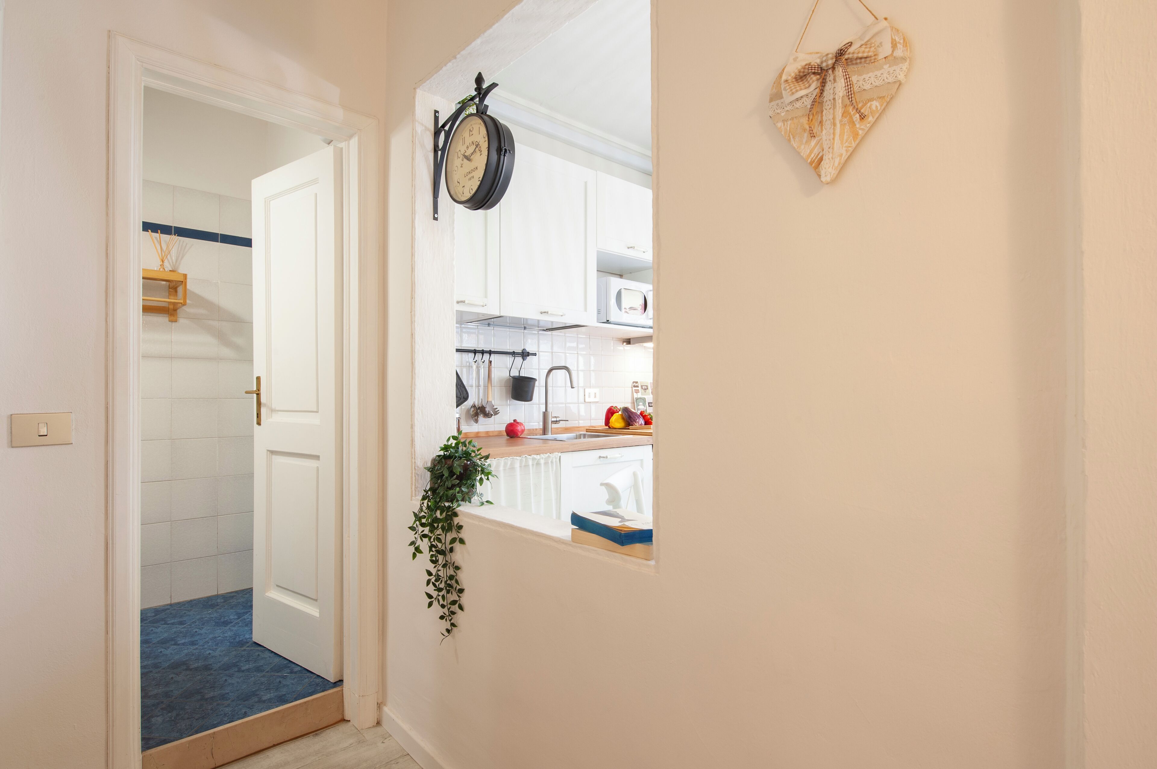 A hallway leading to a kitchen with a white door, a hanging clock, and a heart-shaped decoration.