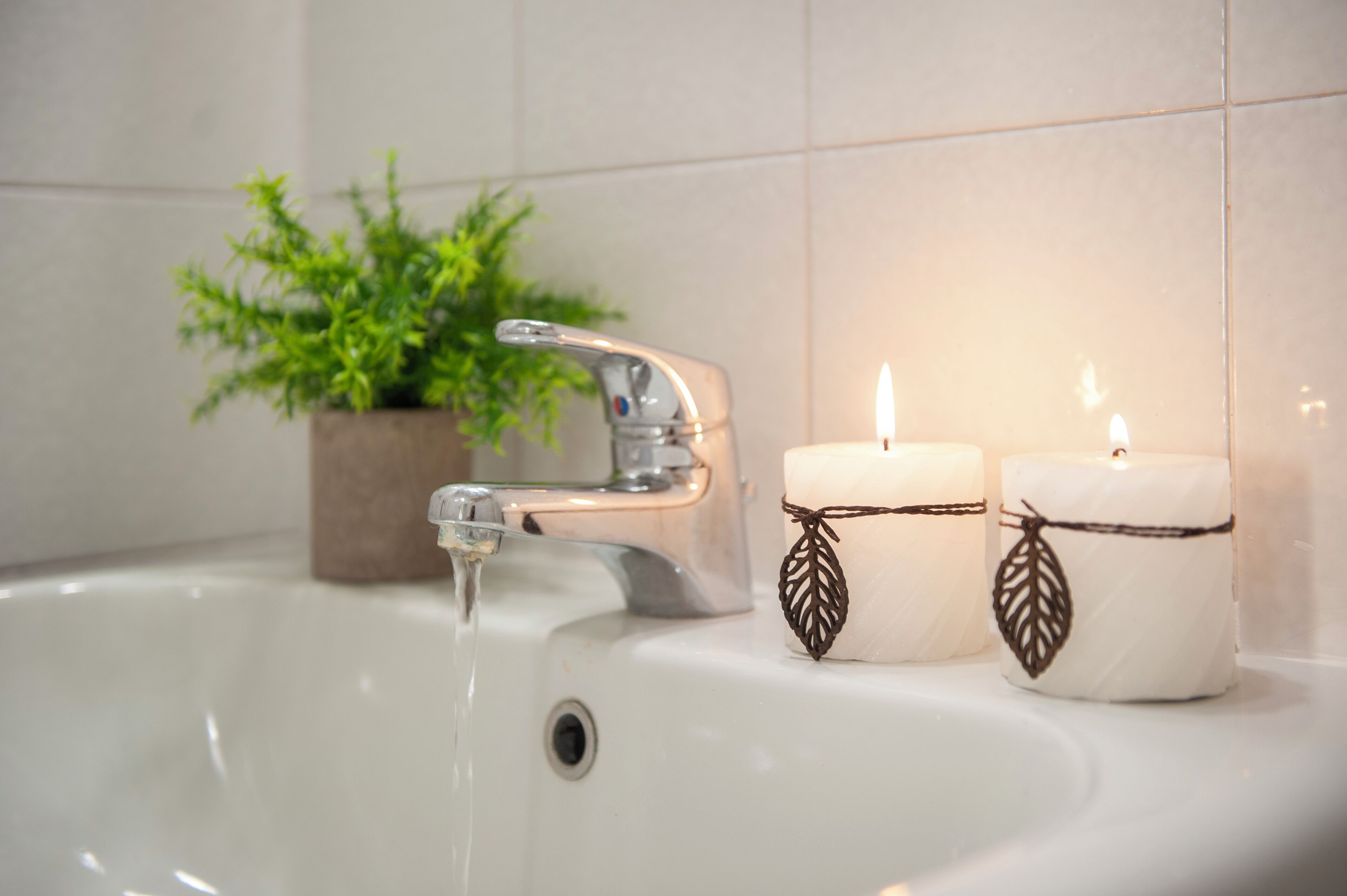 Un lavabo dans une salle de bain avec l’eau qui coule, deux bougies décorées de motifs de feuilles et, au fond, une plante en pot.