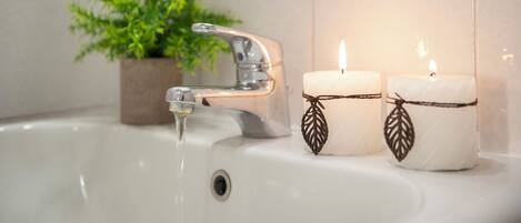 Un lavabo dans une salle de bain avec l’eau qui coule, deux bougies décorées de motifs de feuilles et, au fond, une plante en pot.