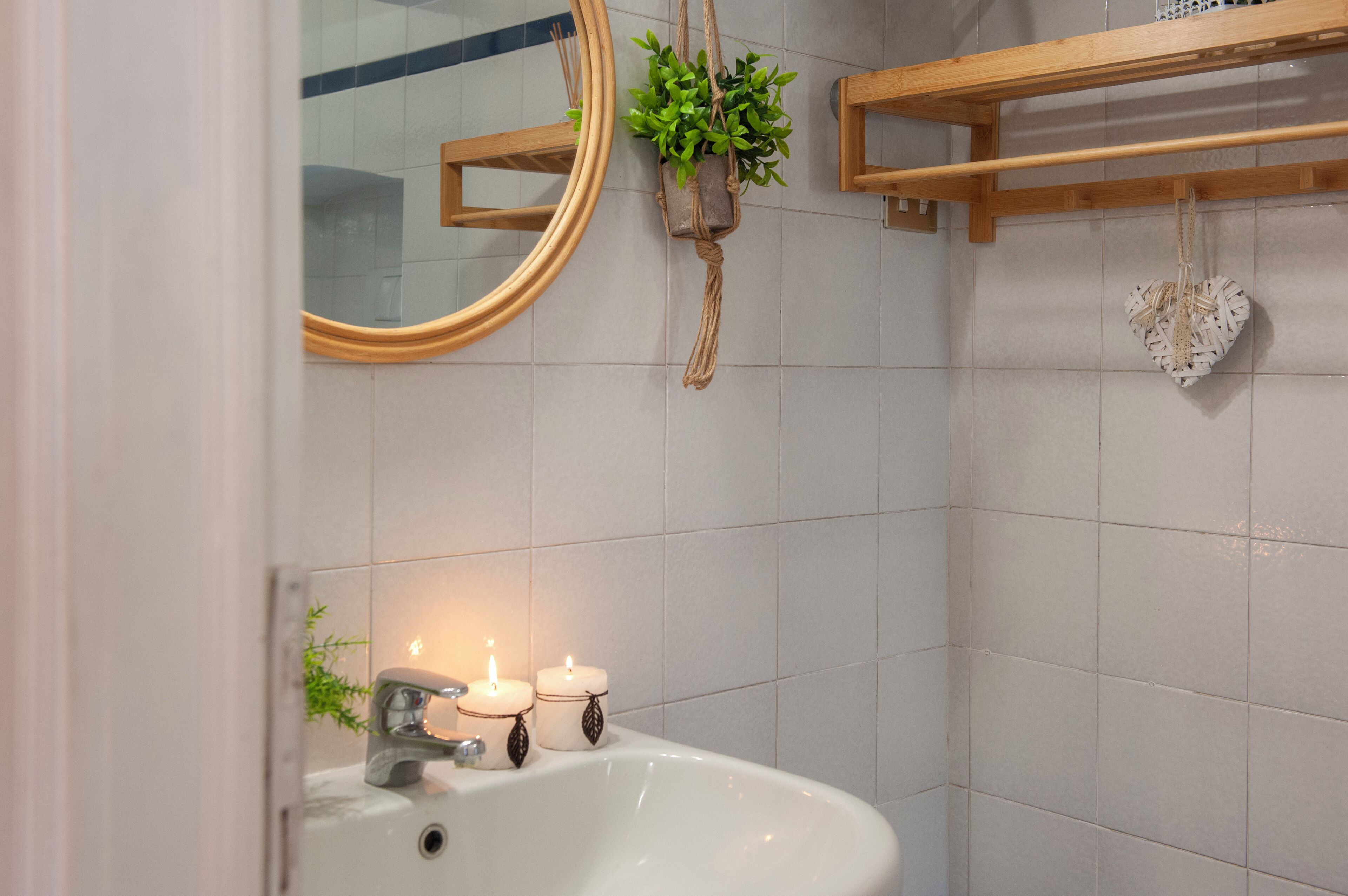 A bathroom with a white sink, two lit candles, a mirror, a wooden shelf with a plant, and a heart-shaped decoration.