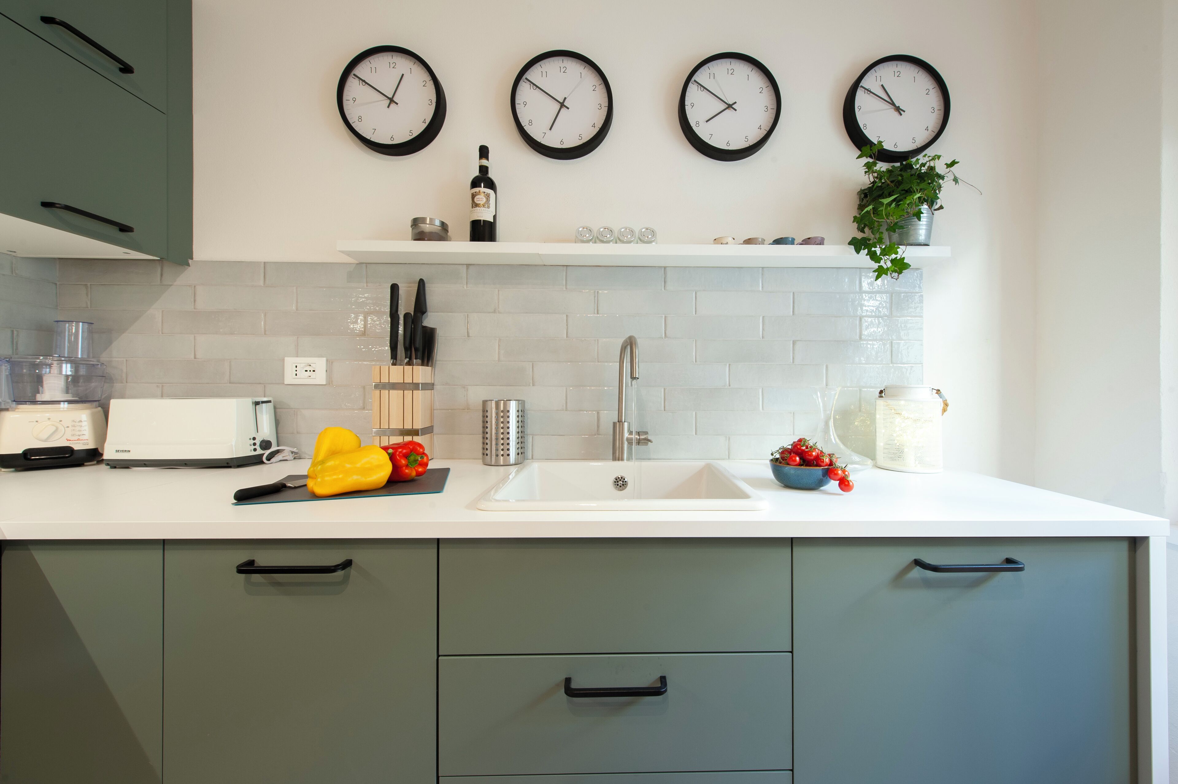 Una cocina con encimera blanca, armarios verdes, un lavabo blanco y tres relojes de pared.