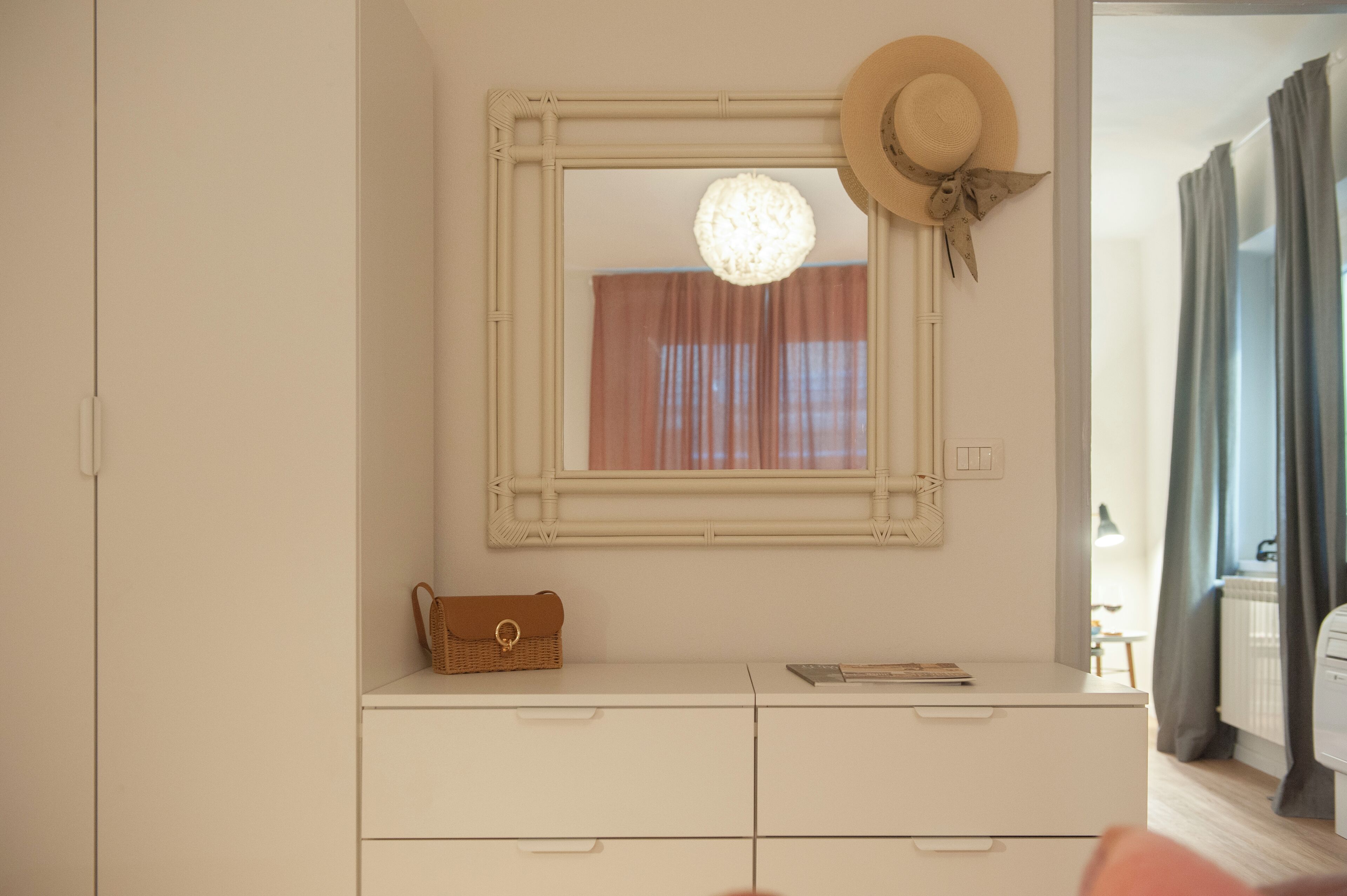 A room with a white dresser, a mirror with a woven frame, a wicker handbag, and a hat on the wall.