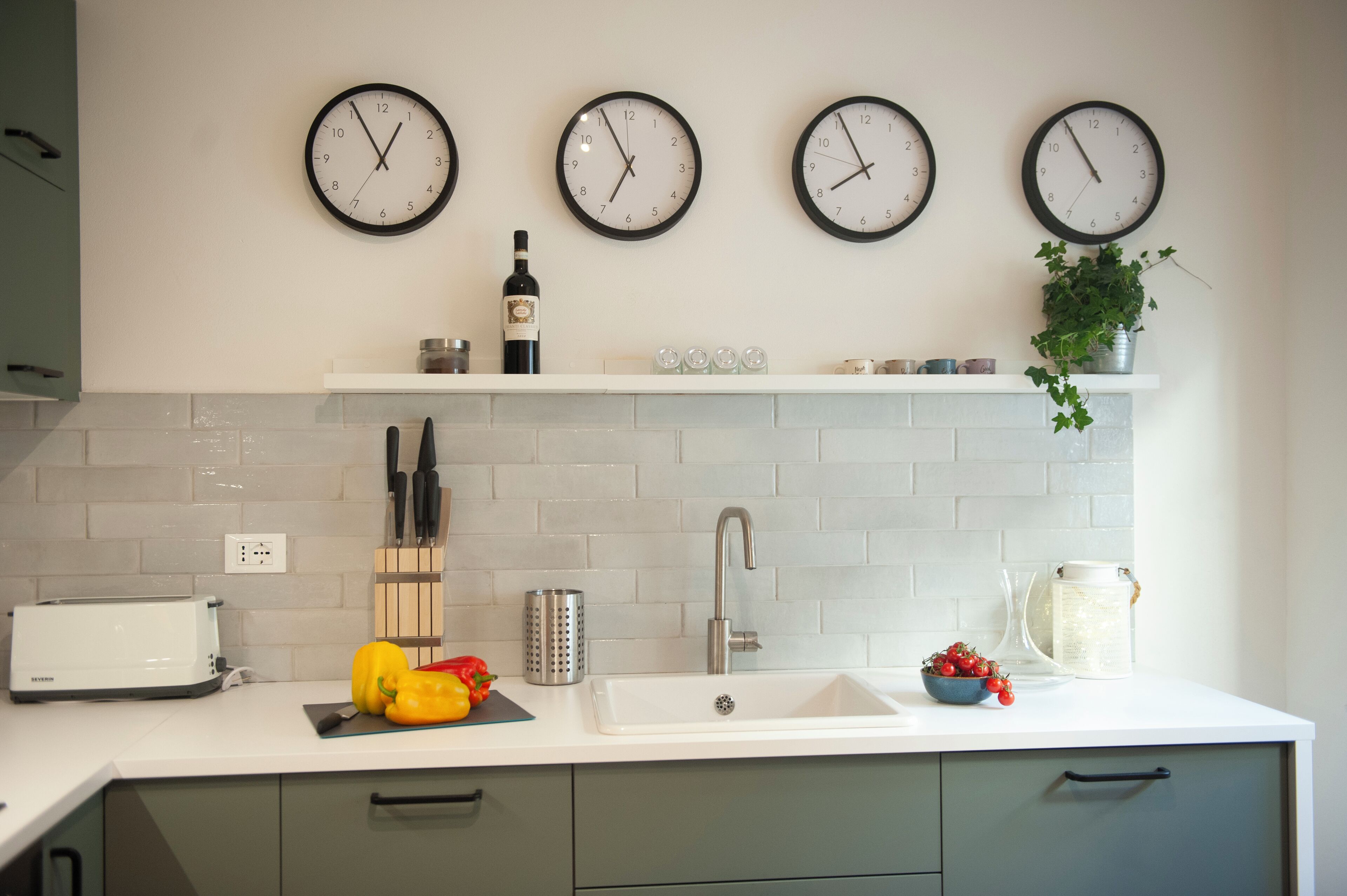 Una cocina con cuatro relojes en la pared, un tostador, un lavabo y un estante con una botella y una planta.