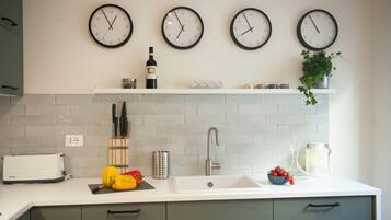 Una cocina con cuatro relojes en la pared, un tostador, un lavabo y un estante con una botella y una planta.