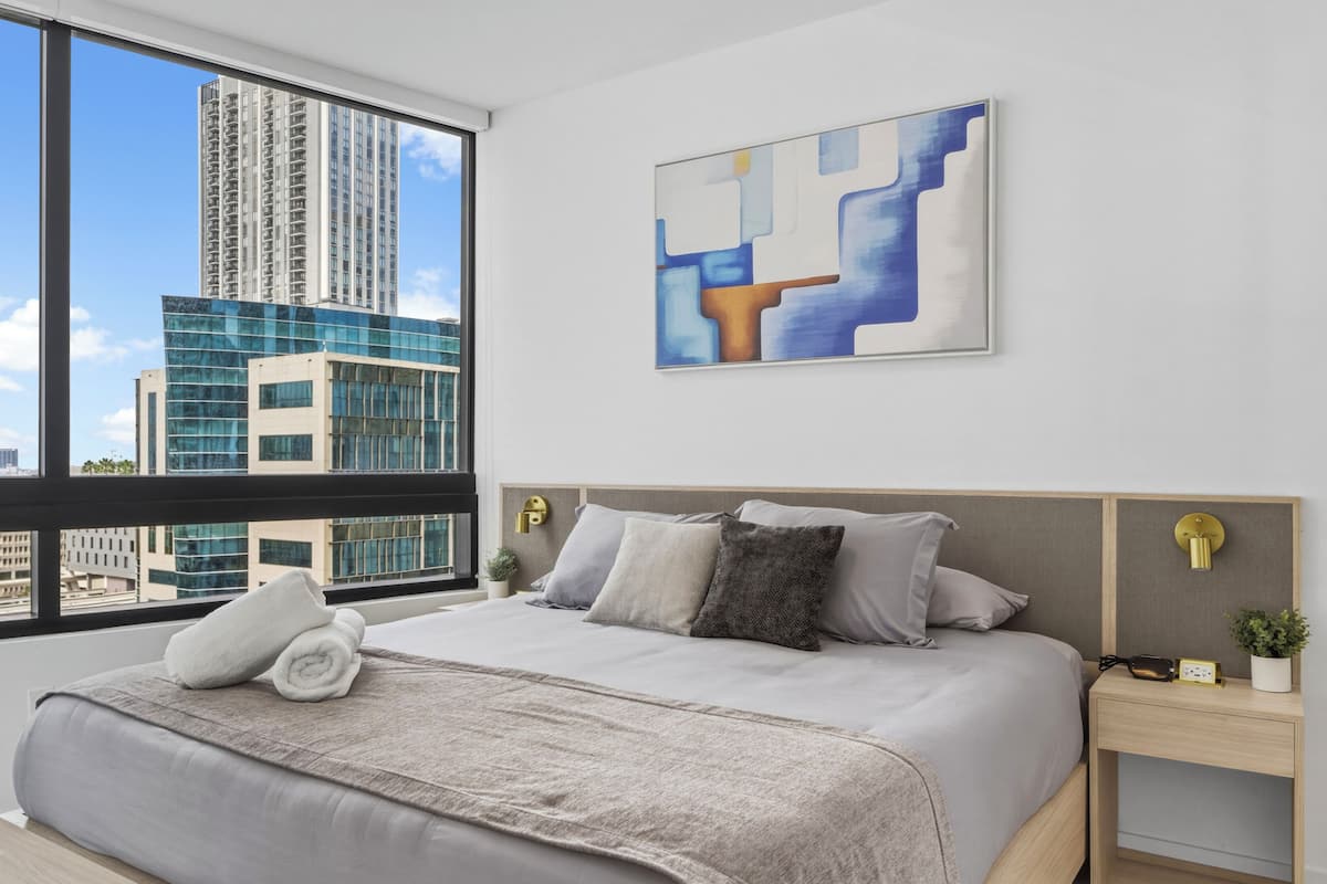 Una habitación de hotel moderna con una cama grande, vistas a edificios altos y un cuadro abstracto contemporáneo.