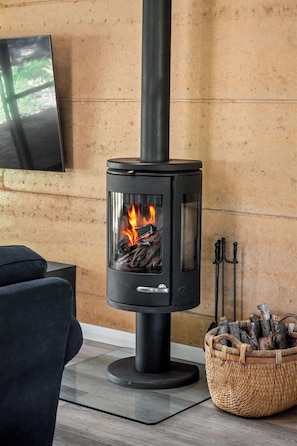 A wood-burning stove with a glass door, a black chimney, and a basket of firewood on the floor. - Waterlily Retreat—Bushland Serenity near the Beach (Eagle Bay)