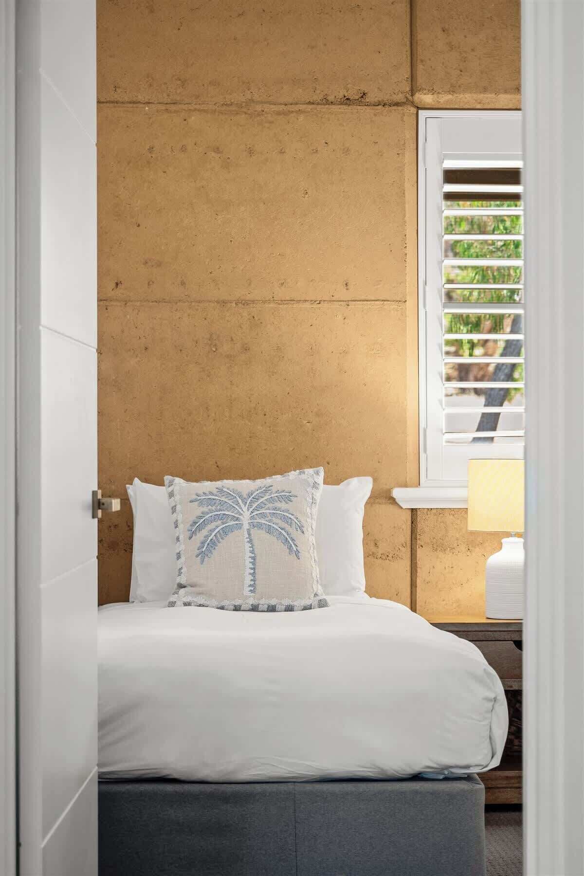 Une chambre à coucher avec un lit, un oreiller orné d’un motif de palmier, une fenêtre avec des stores et une table de chevet avec une lampe.