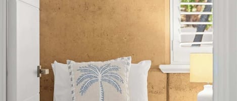 Une chambre à coucher avec un lit, un oreiller orné d’un motif de palmier, une fenêtre avec des stores et une table de chevet avec une lampe.