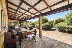 An outdoor patio with a wooden table and benches, a grill, and a covered area with a corrugated roof.
