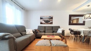 Uma sala de estar moderna com um sofá cinzento, um banquinho de madeira, uma mesa de jantar com cadeiras e um quadro na parede.