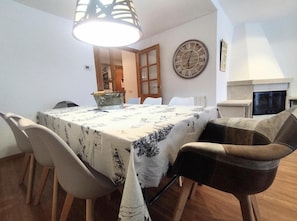 A dining area with a table covered in a patterned tablecloth, surrounded by chairs. A clock is mounted on the wall, and a fireplace is visible in the background. - Casa Arguis Country House with Mountain Views, Wi-Fi, and Air Conditioning (Eriste)