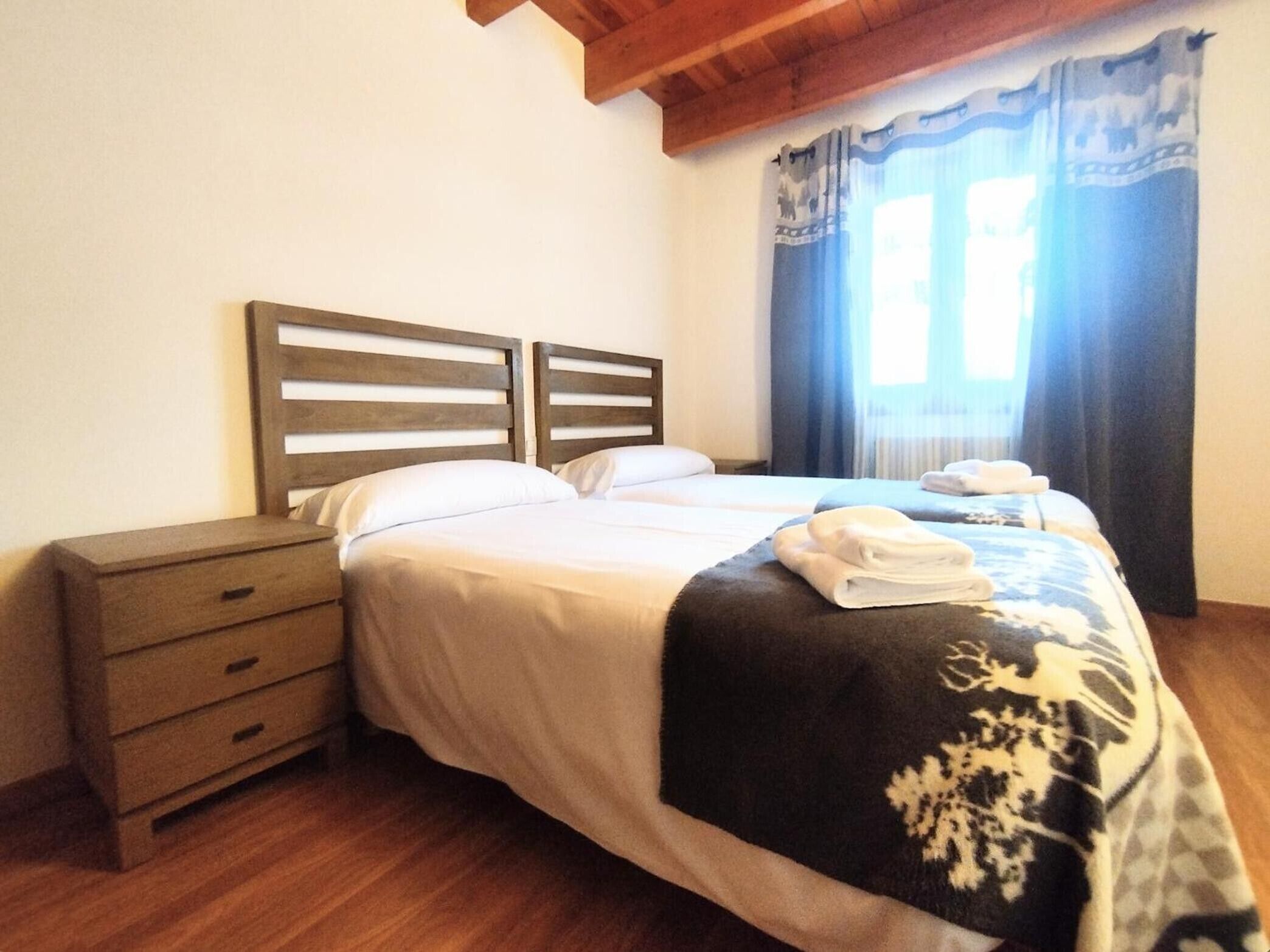 Una cama doble con cabecera, una mesita de noche con cajones y una ventana con cortinas.