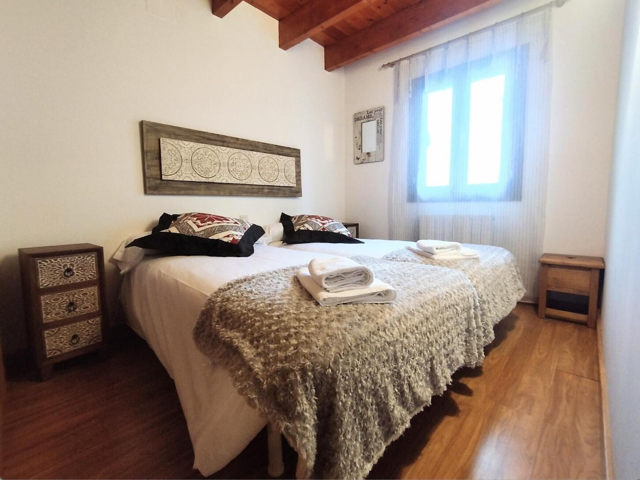 Una cama doble con cabecera estampada, una pieza decorativa en la pared superior y una ventana con cortinas traslúcidas.