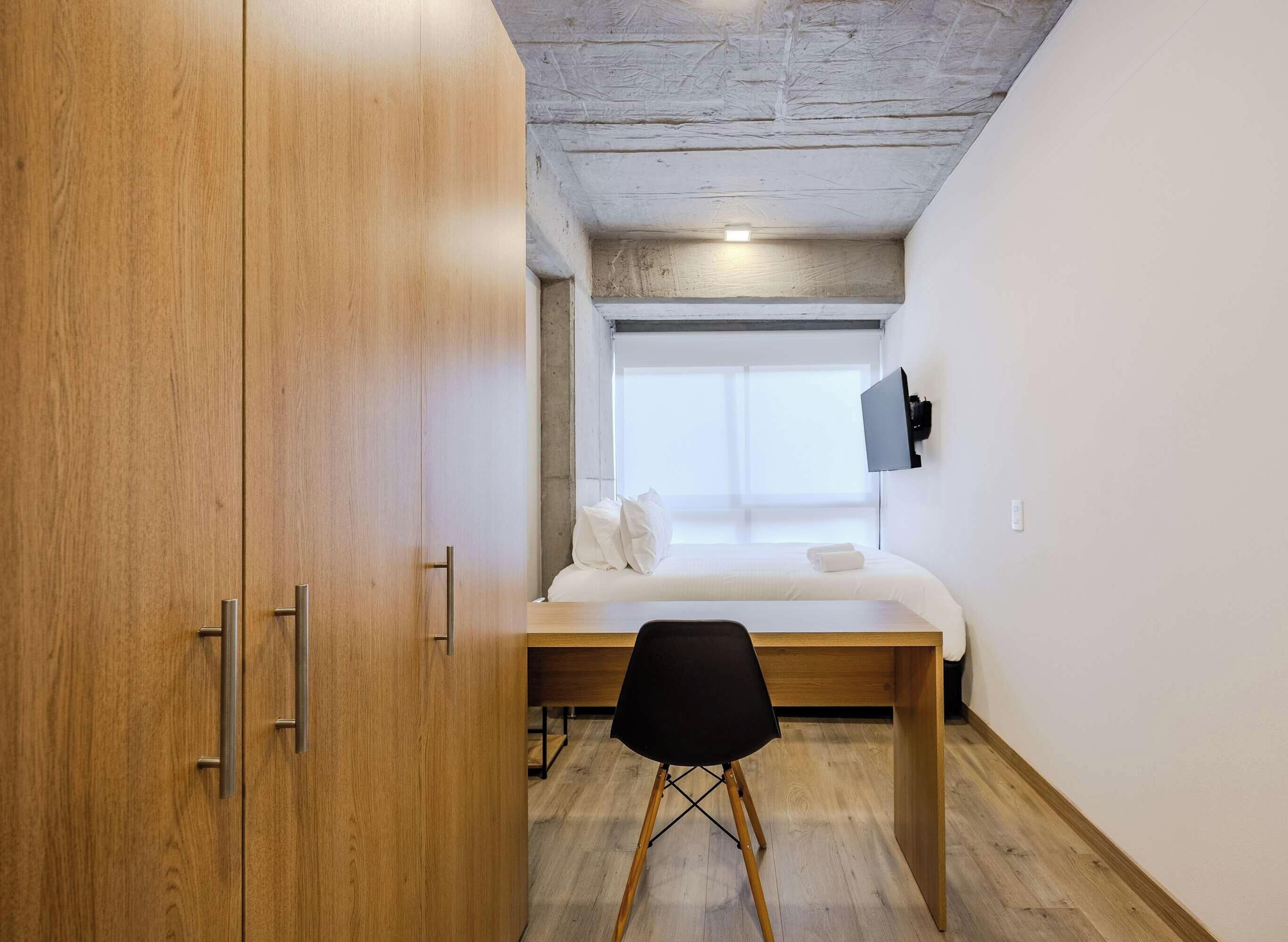 Un dormitorio compacto con un armario de madera, una cama con ropa blanca, un escritorio pequeño con una silla y un televisor montado en la pared.