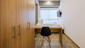 Ein gemütliches Schlafzimmer mit einem Holzschrank, einem Bett mit weißer Bettwäsche, einem kleinen Schreibtisch mit Stuhl und einem an der Wand befestigten Fernseher.