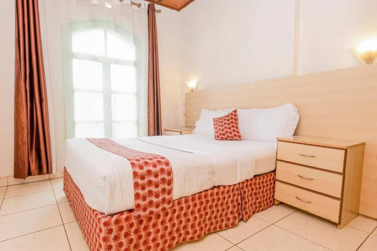 Una cama bien hecha con una colcha estampada y almohadas, una mesita de noche de madera con cajones y una ventana con cortinas.