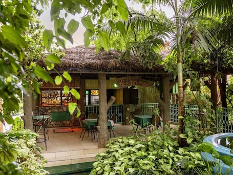 An outdoor restaurant with a thatched roof, surrounded by lush greenery and a variety of plants.
