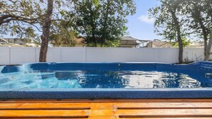Una piscina con plataforma de madera, rodeada por una valla blanca y árboles.