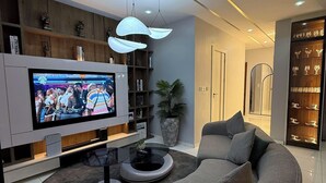 A modern living room with a large flat-screen TV, a curved grey sofa, a coffee table, and a bookshelf. - Osaka Apartment by J's Luxury (Benin City)