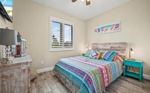 A bedroom with a wooden floor, a bed with a colorful blanket, a nightstand, a lamp, a dresser, and a window with blinds. - Ibis Landing (Fort Myers Beach)