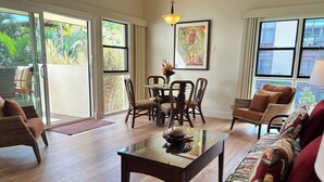 A spacious living room with a glass coffee table, wicker furniture, and a view of a lush garden through large windows. - New Remodel Beachfront Maalaea Condo w/ AC (Wailuku)
