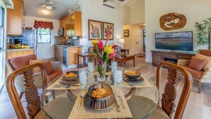 A dining area with a glass table, chairs with pineapple designs, a vase with flowers, and a kitchen in the background. - New Remodel Beachfront Maalaea Condo w/ AC (Wailuku)