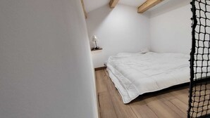 A small, white-walled room with a wooden floor, a single bed, a small table with a lamp, and a netted window. - Bright and spacious apartment in house located between sea and mountains (Nice)