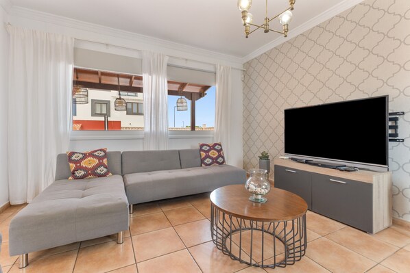 Un salon avec un canapé gris, une table basse ronde, un téléviseur posé sur un meuble et un mur orné d’un motif.
