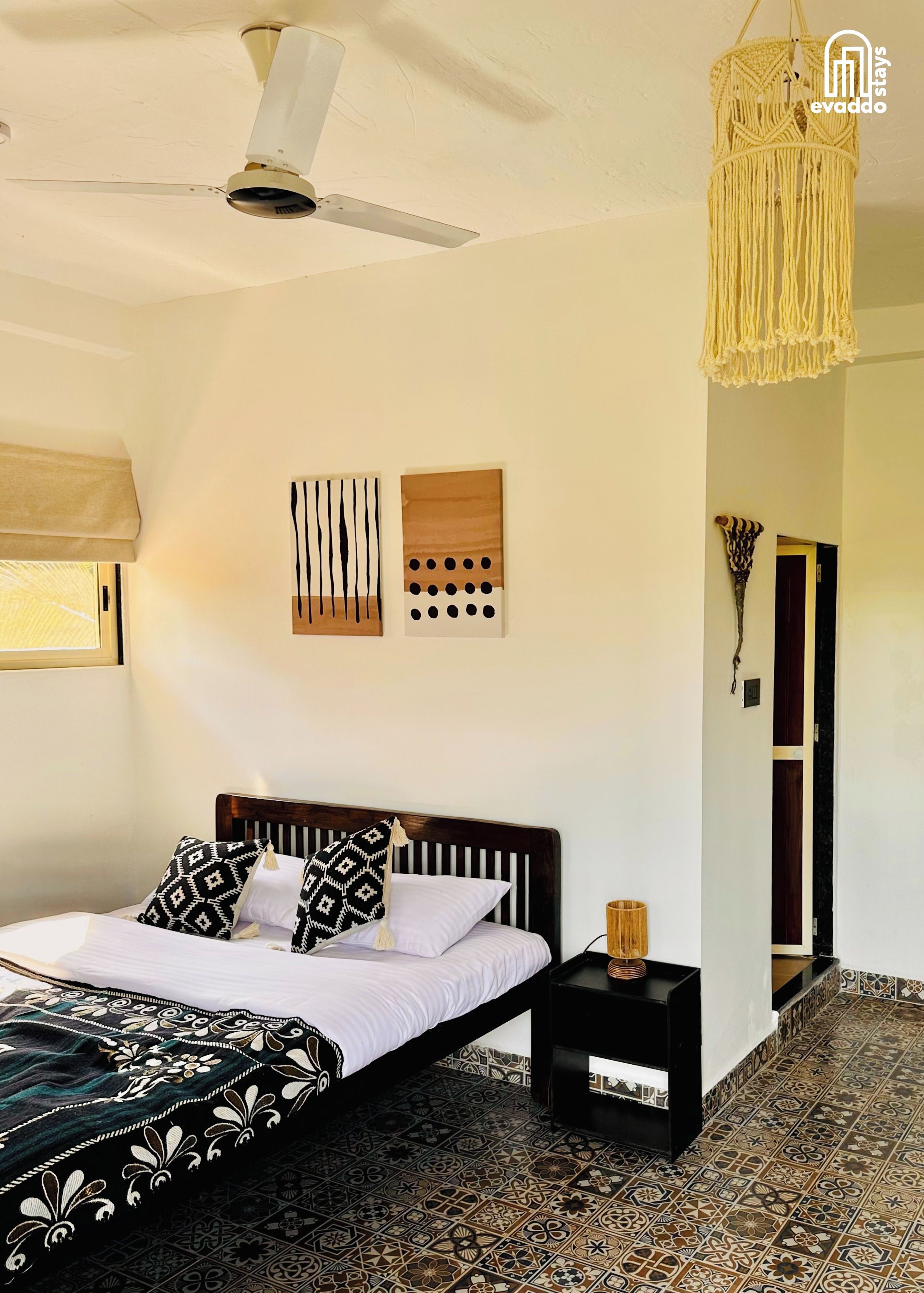 Un dormitorio con cama, mesita de noche, lámpara colgante de macramé y una obra de arte en la pared.