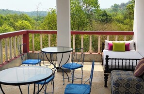 A balcony with a glass table and blue chairs, a bed with patterned bedding, and a view of a green landscape. - Bhumika by evaddo (Ashvem)