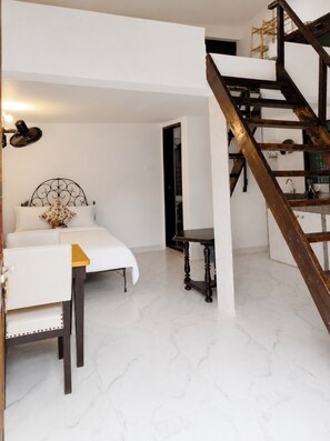A bedroom with a bed, a bedside table, a lamp, a wooden staircase, and a white wall. - Bhumika by evaddo (Ashvem)