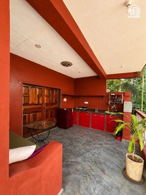 A compact kitchen with red cabinets, a wooden door, and a small dining table. - Bhumika by evaddo stays (Ashvem)