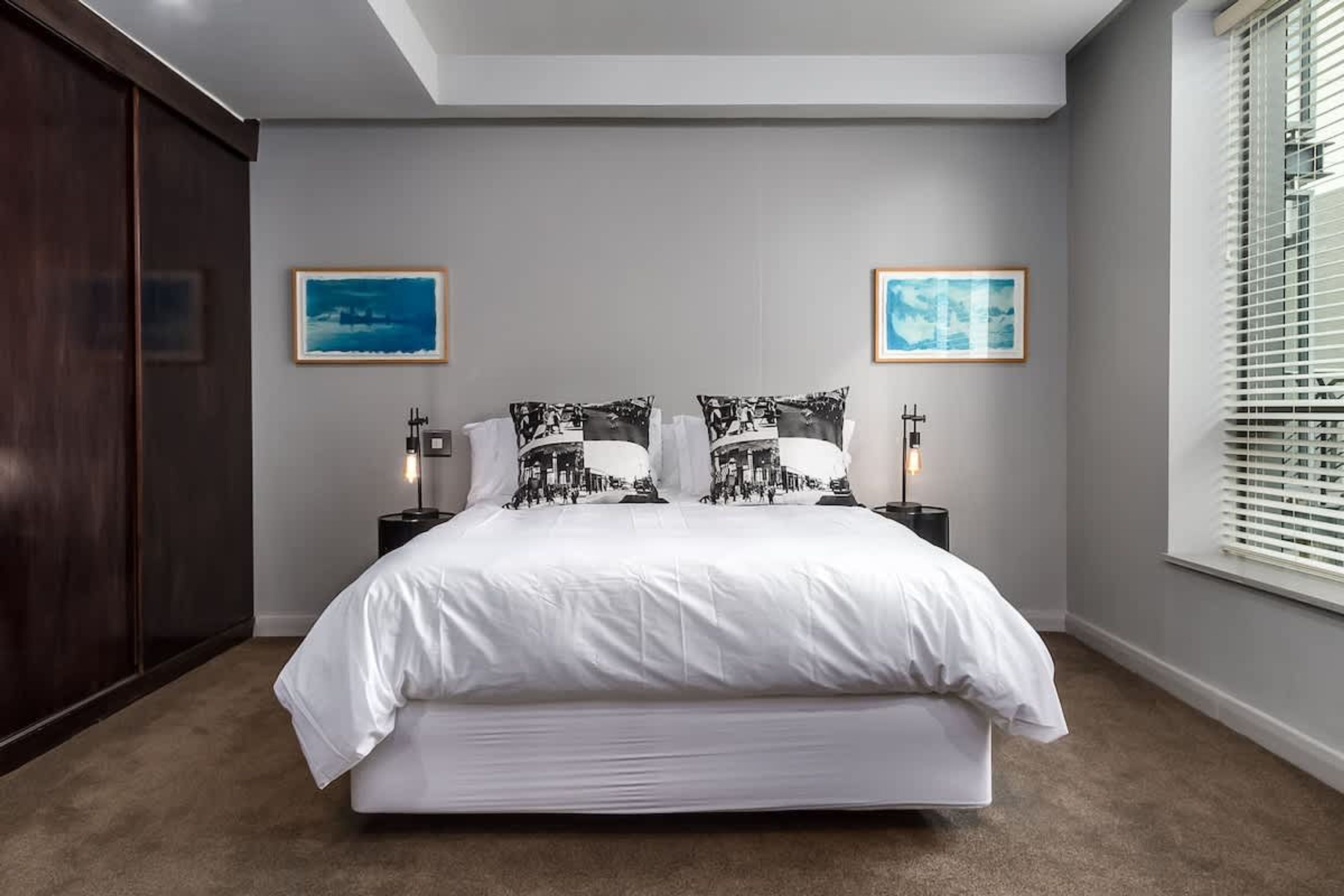 Camera da letto con un grande letto, due comodini con lampade, una finestra con tende e due quadri appesi al muro.